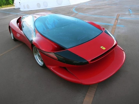 A red Ferrari concept car sits on a paved surface. The car has a large, tinted glass bubble covering the cockpit area. The rear of the car features a wide, flat spoiler and a black accent stripe. The Ferrari logo is visible on the back. In the background, a white tent-like structure is partially visible. The ground has faded blue and orange lines on it.

Provided by @altbot, generated using Gemini