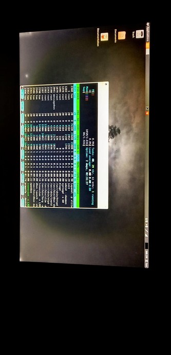 The photograph shows a computer LCD monitor displaying a terminal window with a Bash command-line interface. The terminal window is open and shows a list of processes, with columns for various process details such as user, priority, memory usage, and CPU usage. The terminal window is titled "top" and is running on a Linux system, as indicated by the "Linux" text at the bottom of the screen. The desktop background is a dark image of a cloudy sky with a tree visible in the distance. The top of the screen shows several icons, including "Documentation," "Documents," and "Trash," along with a "Applications" menu. The taskbar on the right side of the screen shows the system time, date, and some system information, including the load average and the number of tasks. The overall color scheme of the screen is dark, with text and icons in various shades of white, green, and blue.